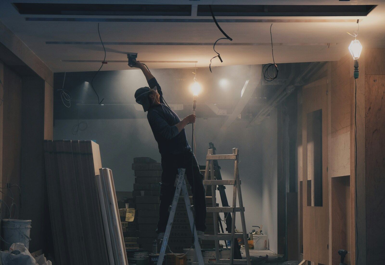 Handwerker auf einer Leiter arbeitet an der Decke eines noch unfertigen, abgedunkelten Innenraums.